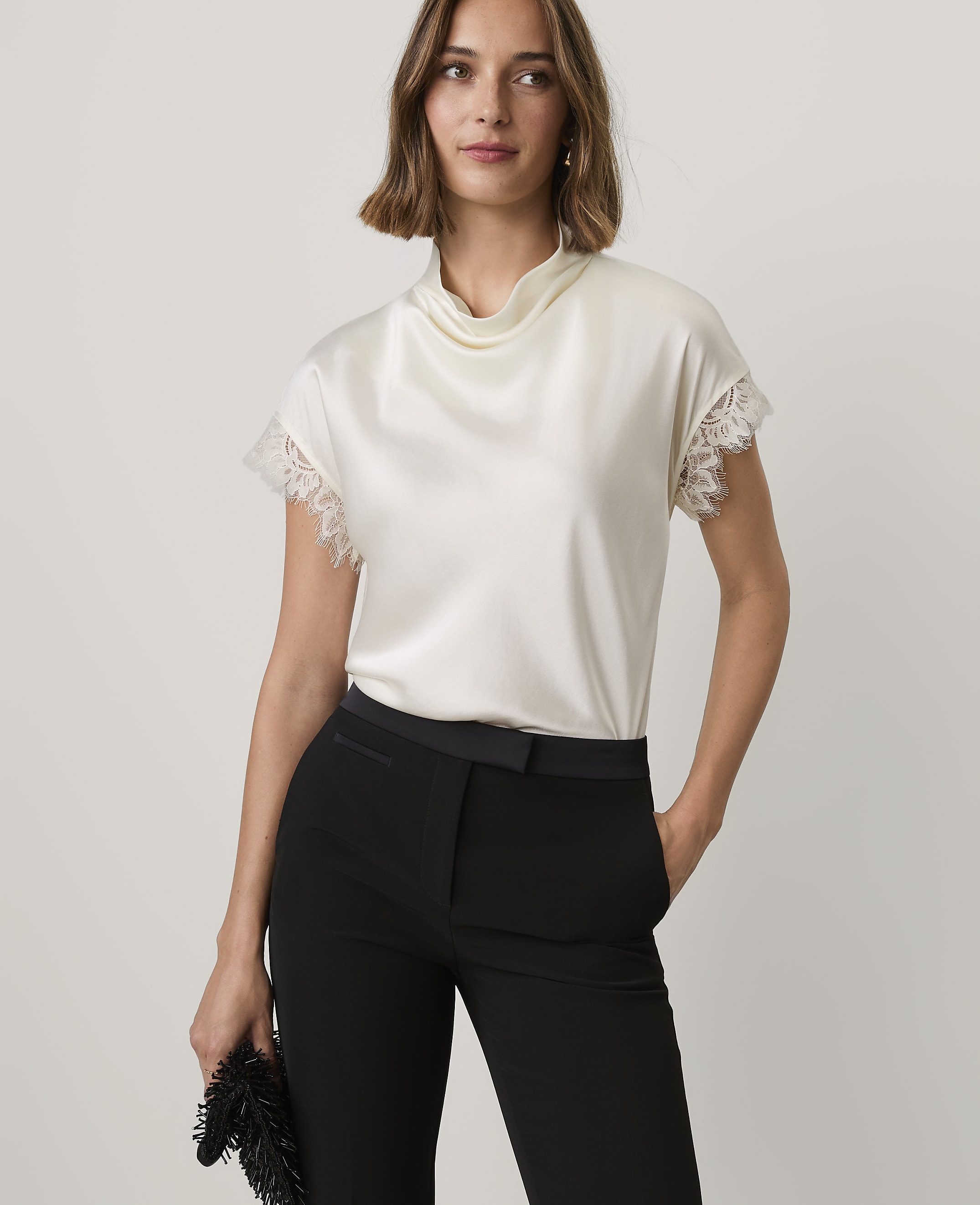 A woman wearing a white satin top and black trousers stands against a plain, light-colored background.