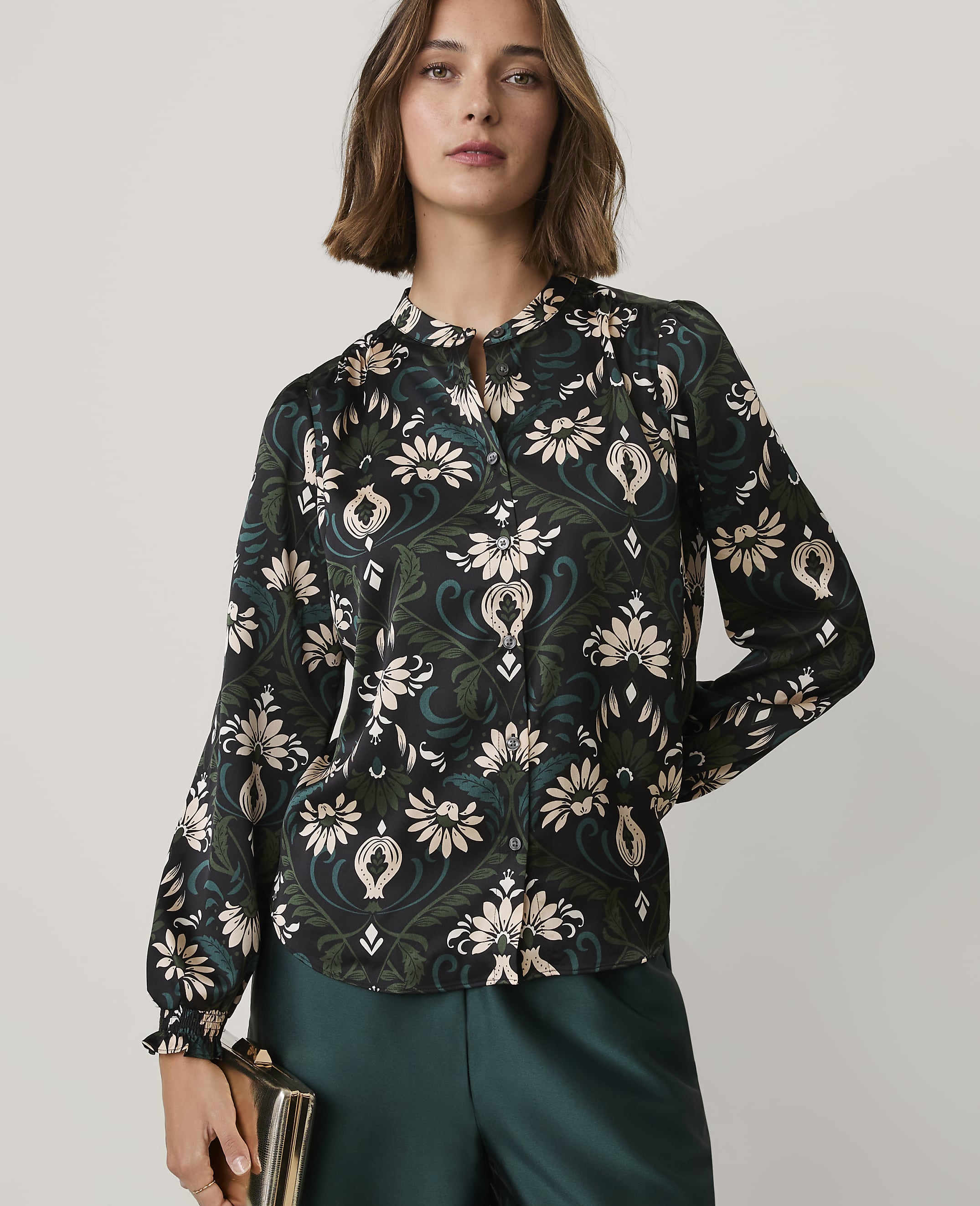 A woman wearing a floral print blouse and dark green trousers stands against a plain, light-colored background.