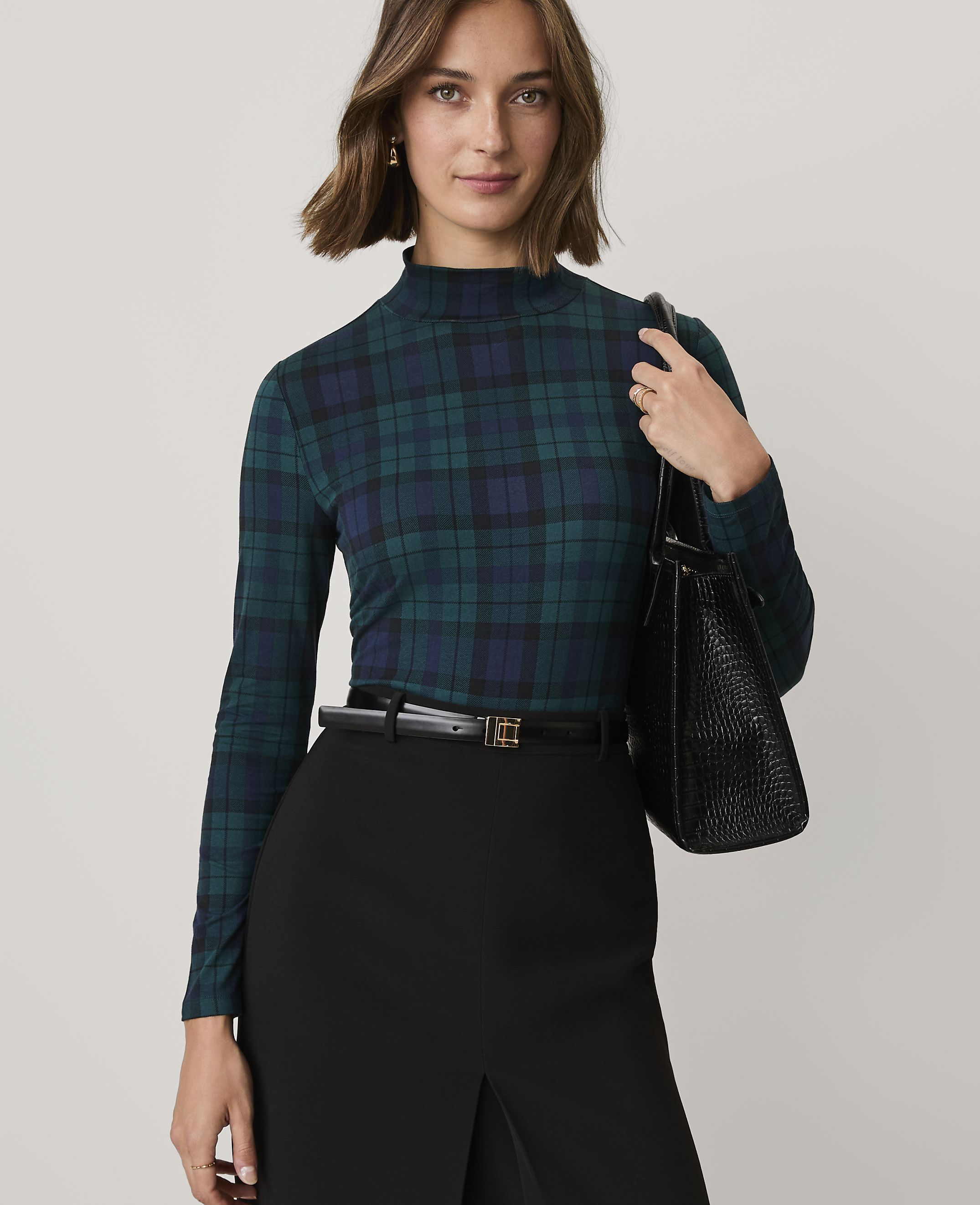 A woman wearing a green plaid turtleneck and black skirt stands against a plain light gray background.