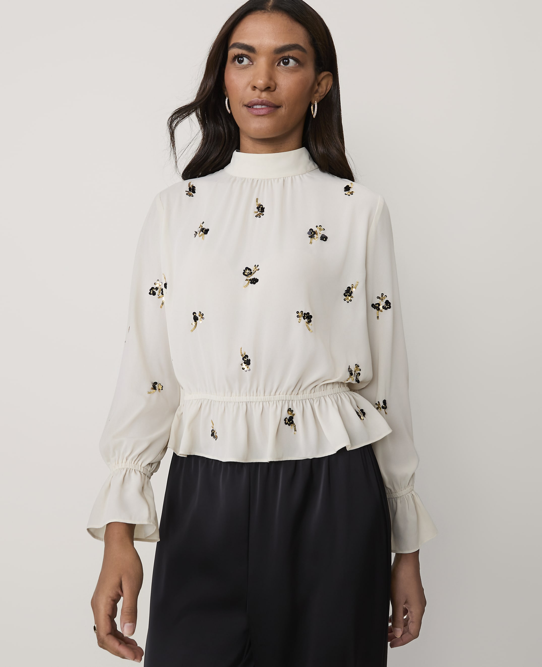 A woman wearing a white floral-embroidered blouse and black trousers stands against a plain light-colored background.