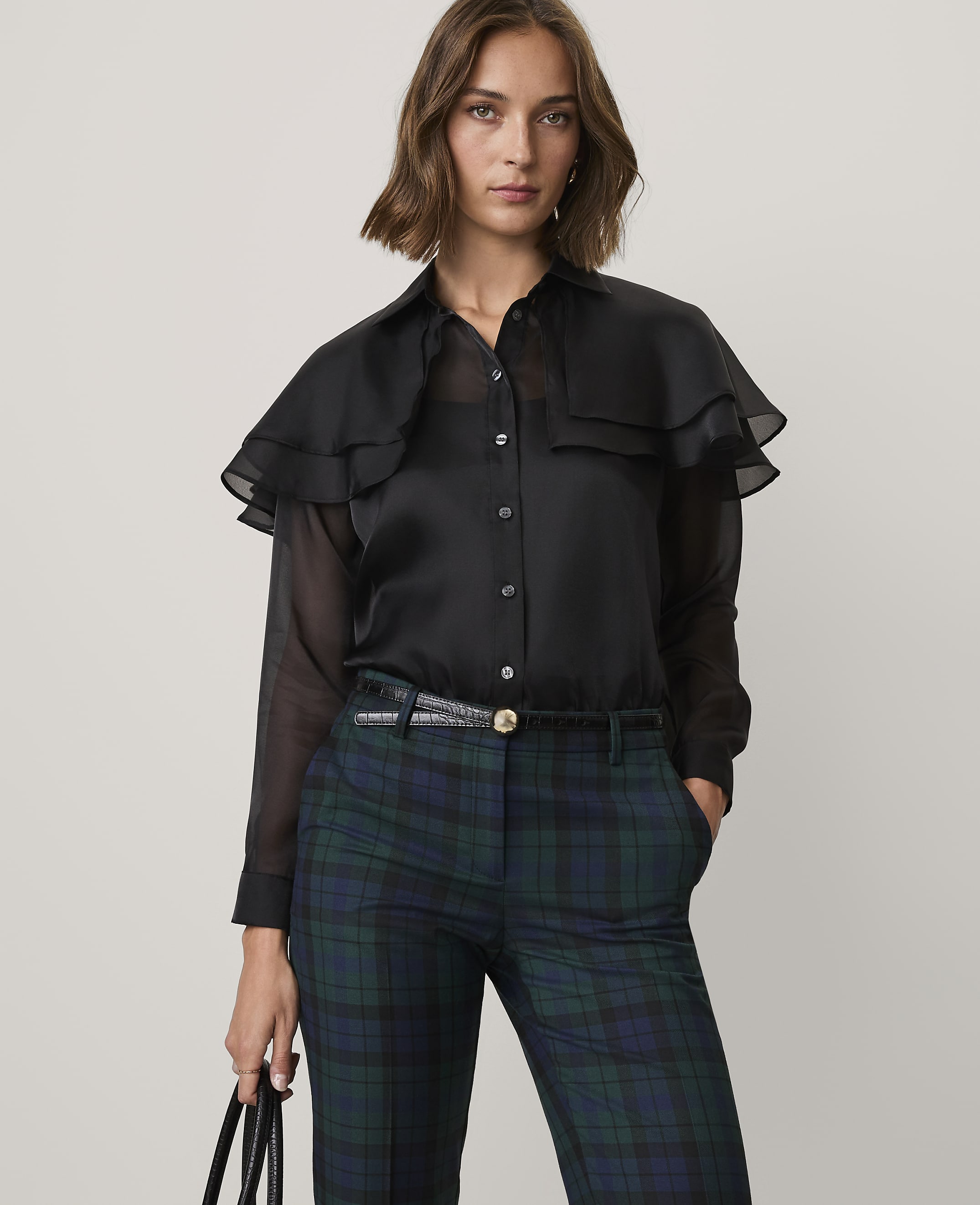 A woman wearing a black ruffled blouse and green plaid trousers stands against a plain light gray background.