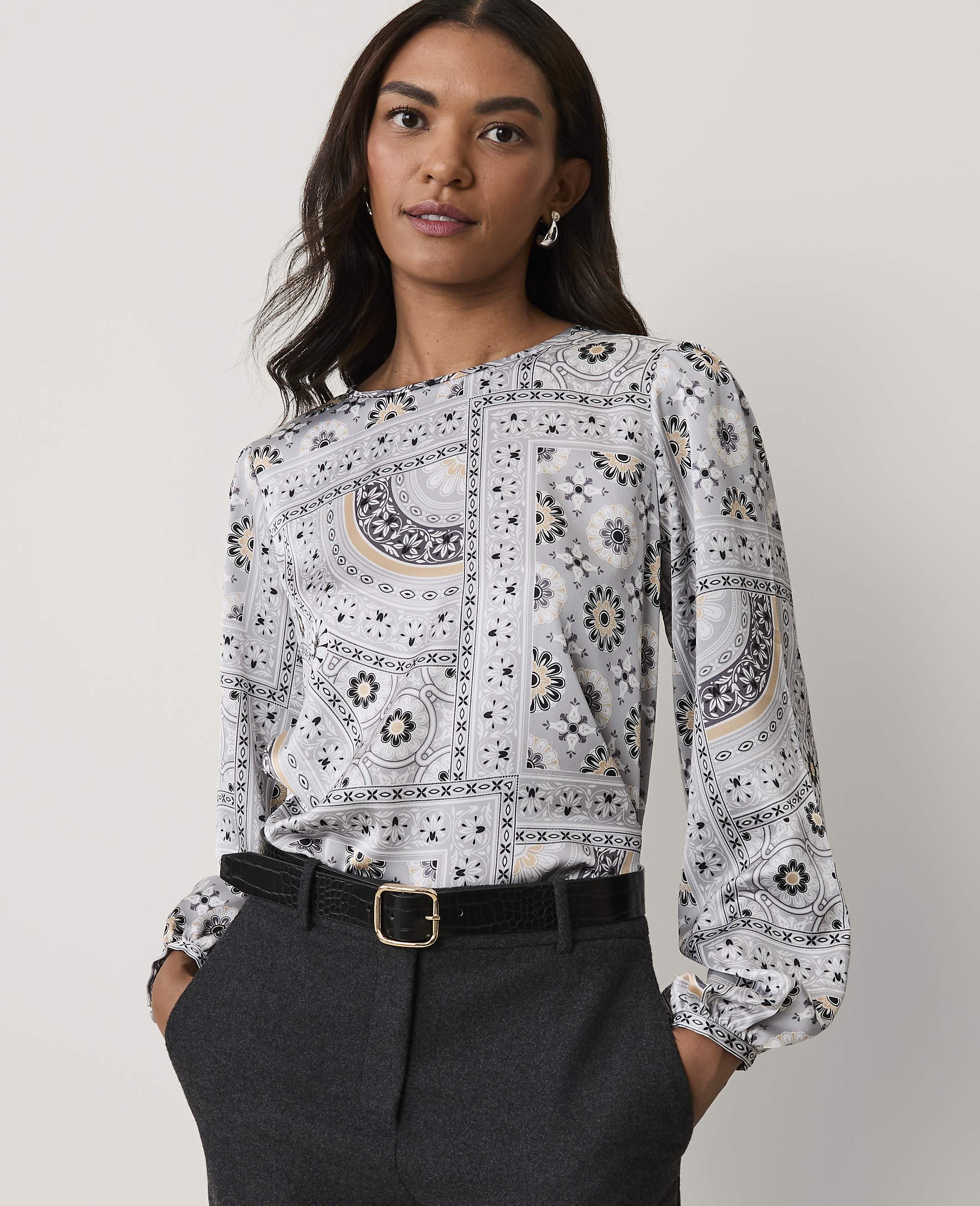 A woman wearing a patterned grey blouse and dark trousers stands against a plain white background.