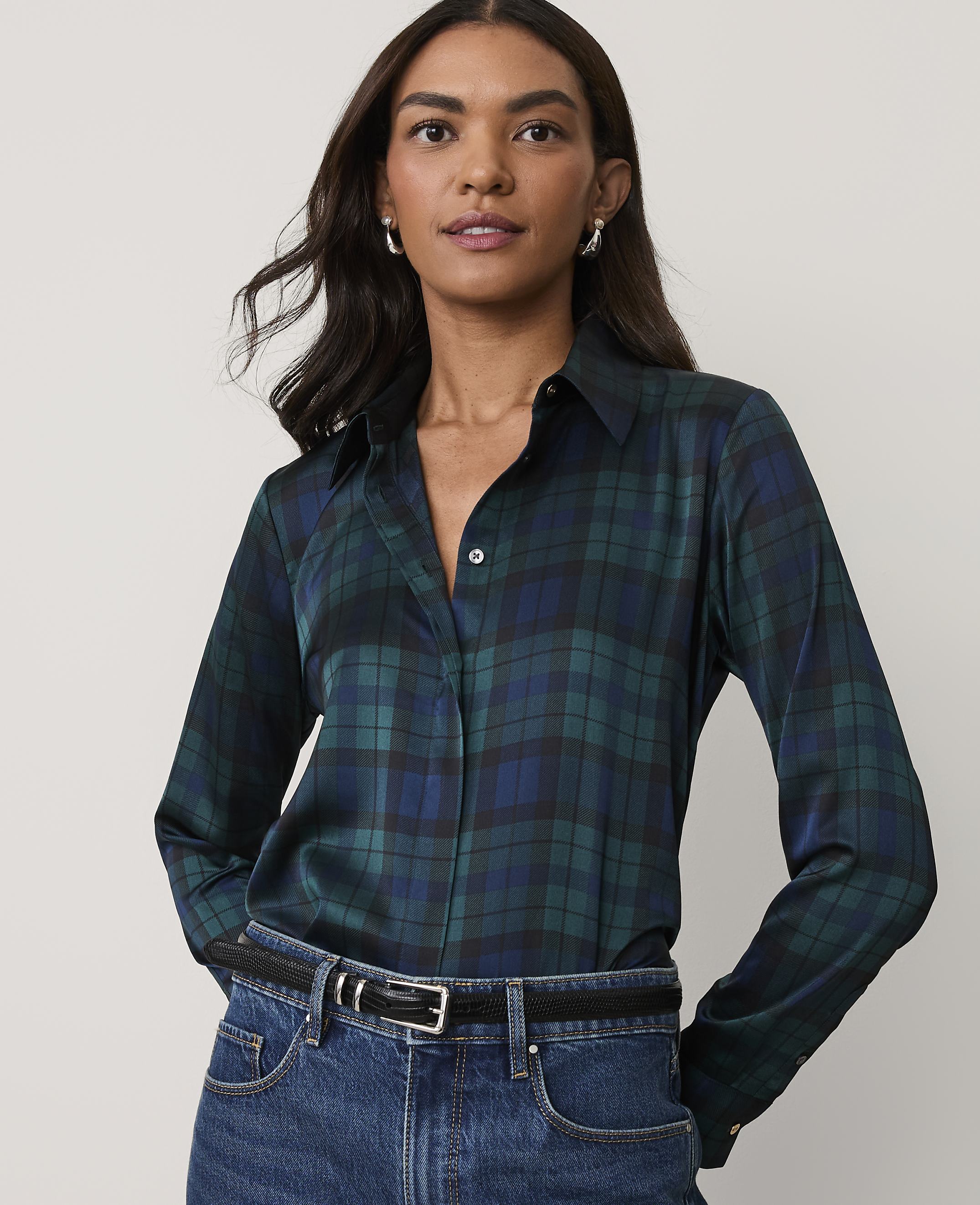 A woman wearing a dark green plaid button-down shirt and blue jeans stands against a plain, light-colored background.