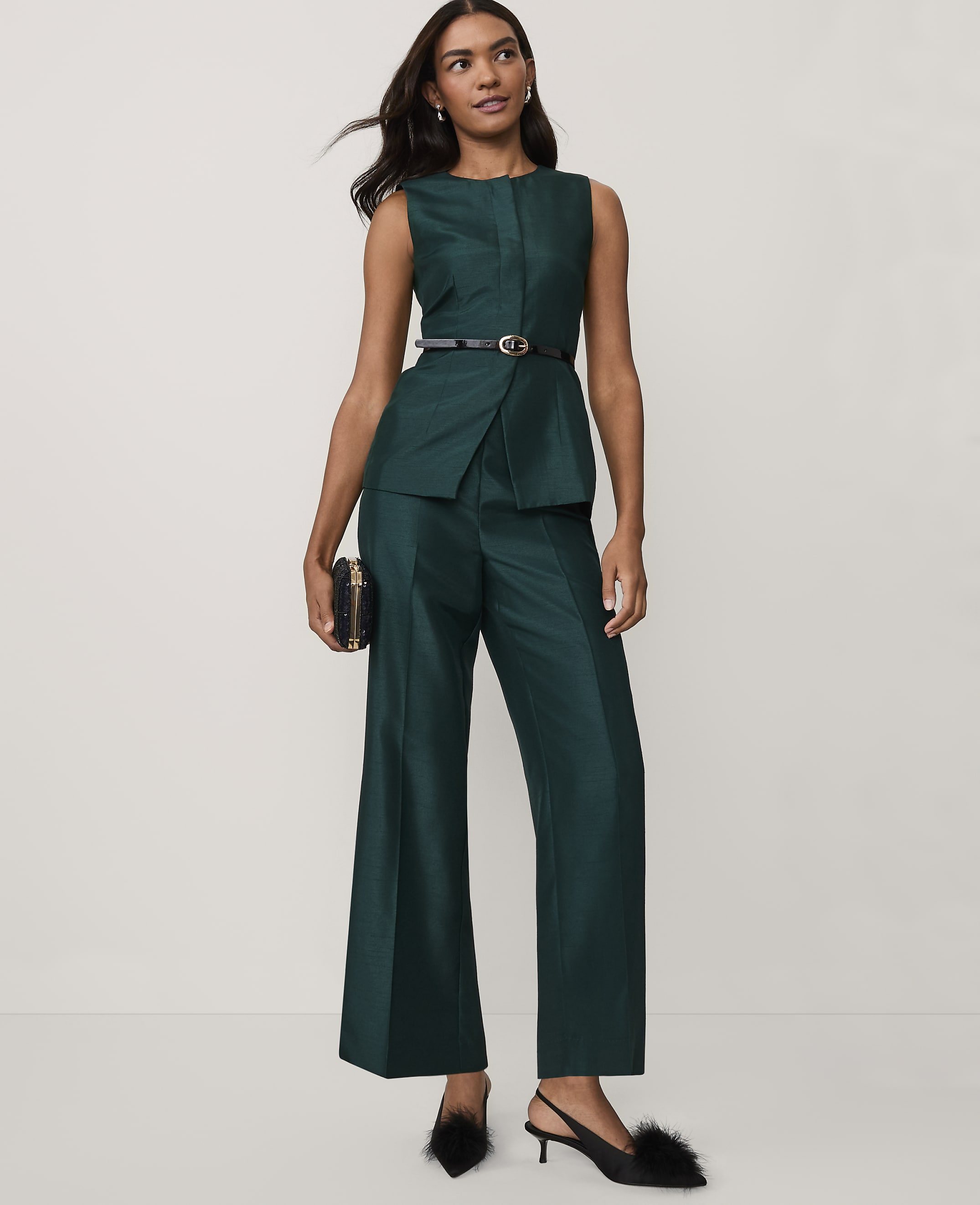 A woman wearing a dark green sleeveless suit and black heels stands against a plain white background.