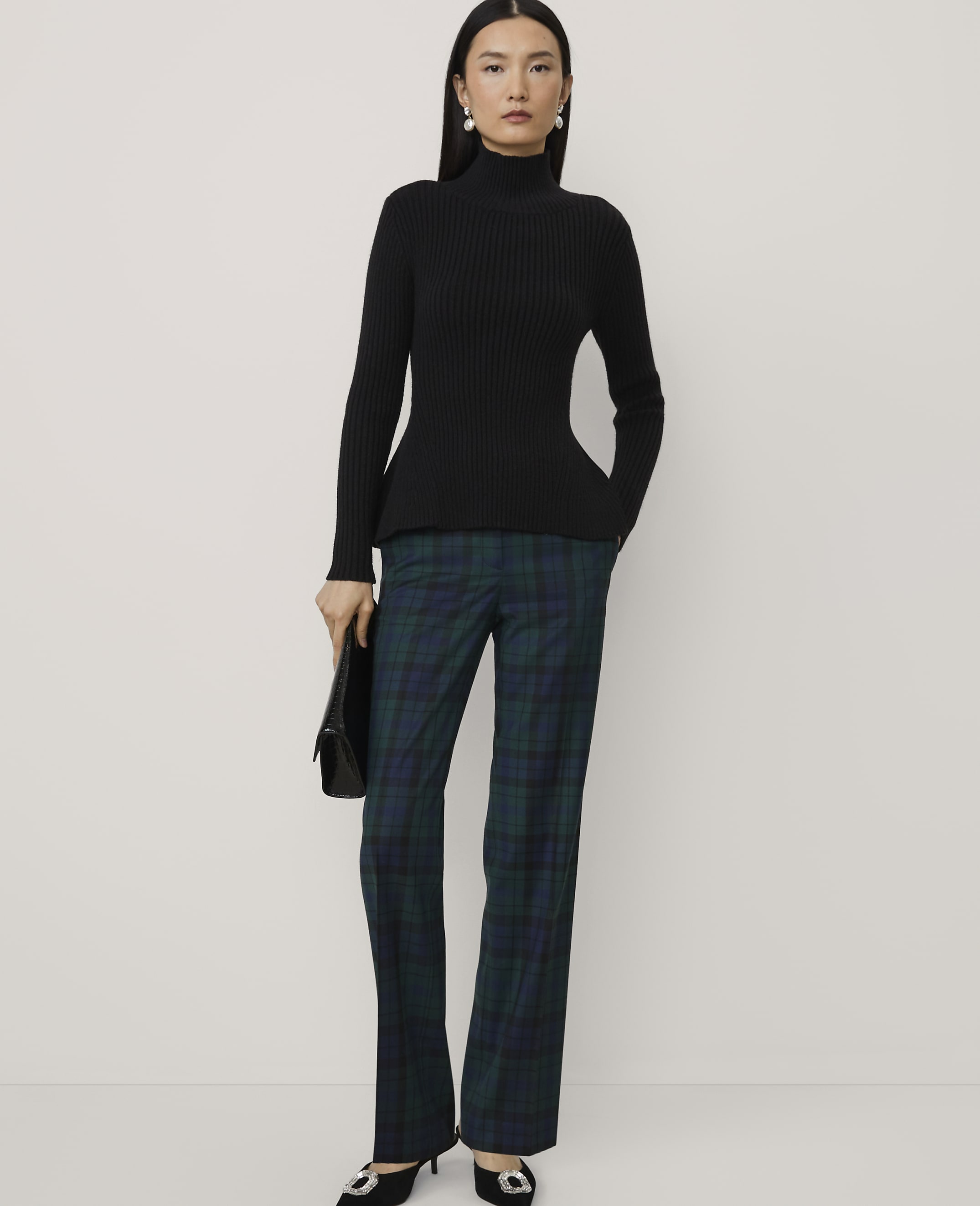 A woman wearing a black ribbed turtleneck and green plaid trousers stands against a plain, light-colored background.