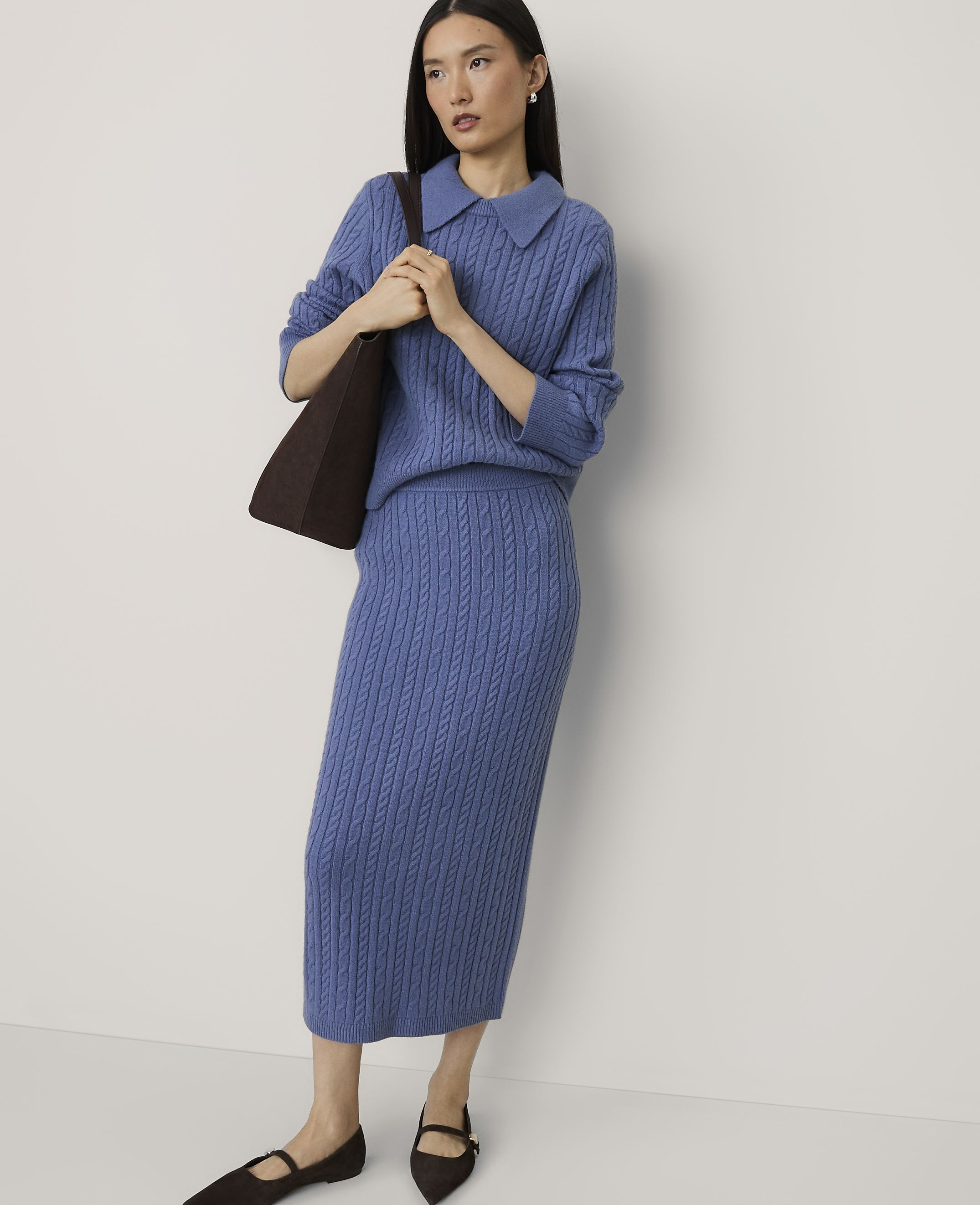 A woman wearing a blue cable-knit sweater and matching skirt stands against a plain white background.