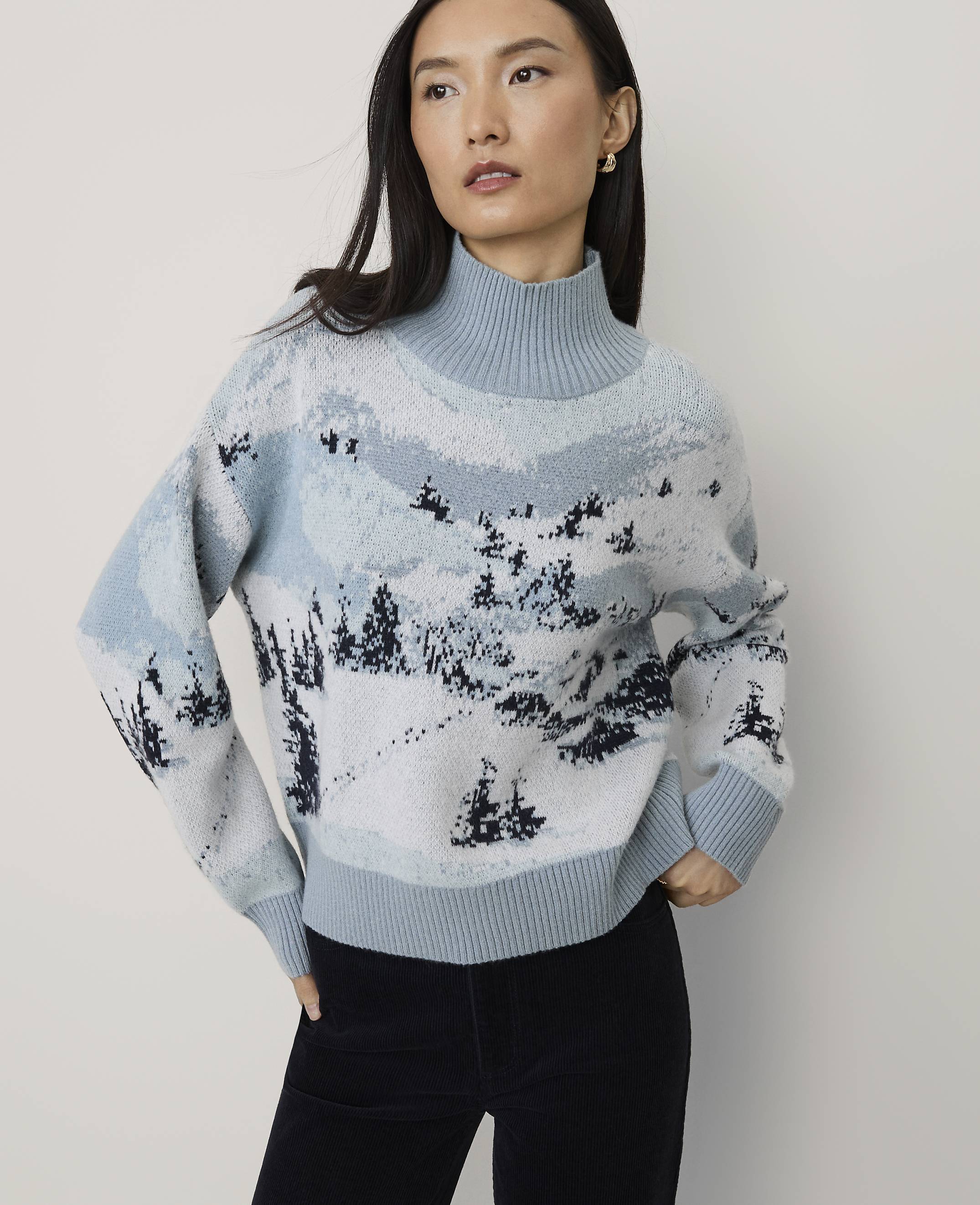 A woman wearing a light blue winter landscape patterned sweater and black pants stands against a plain white background.