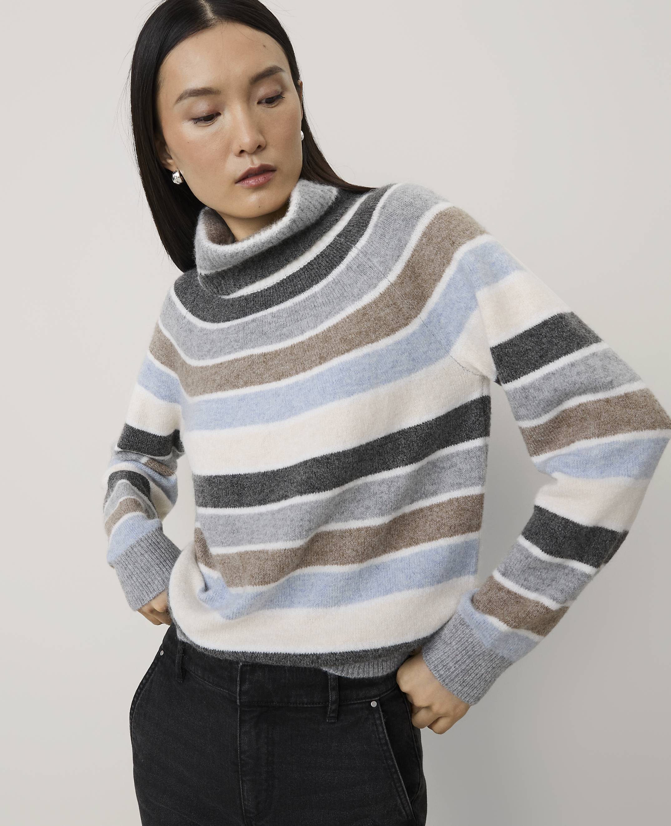 A woman wearing a multi-colored striped turtleneck sweater and black pants stands against a plain, light-colored background.