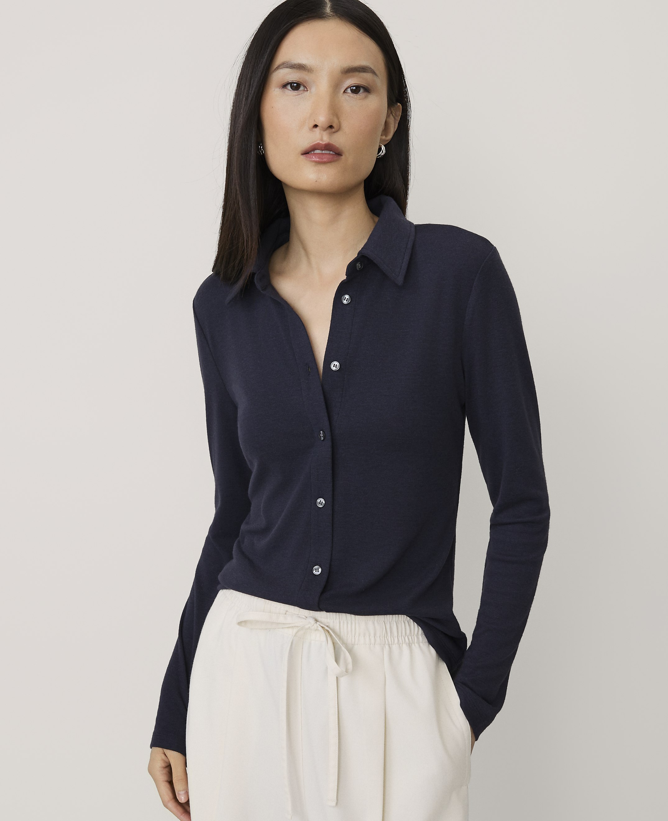 A woman wearing a navy blue long-sleeved collared shirt and white trousers stands against a plain light gray background.
