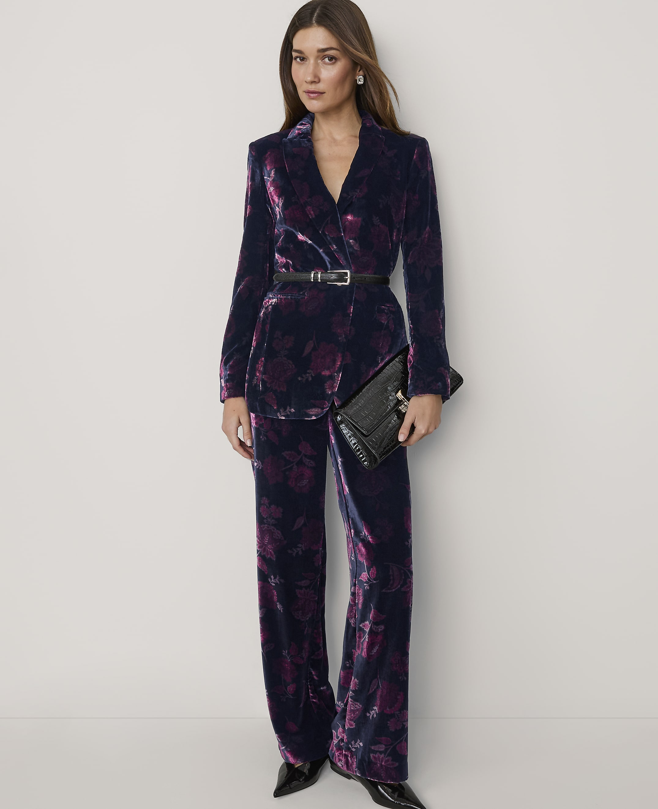 A woman wearing a dark purple velvet floral suit and holding a black clutch stands against a plain white background.