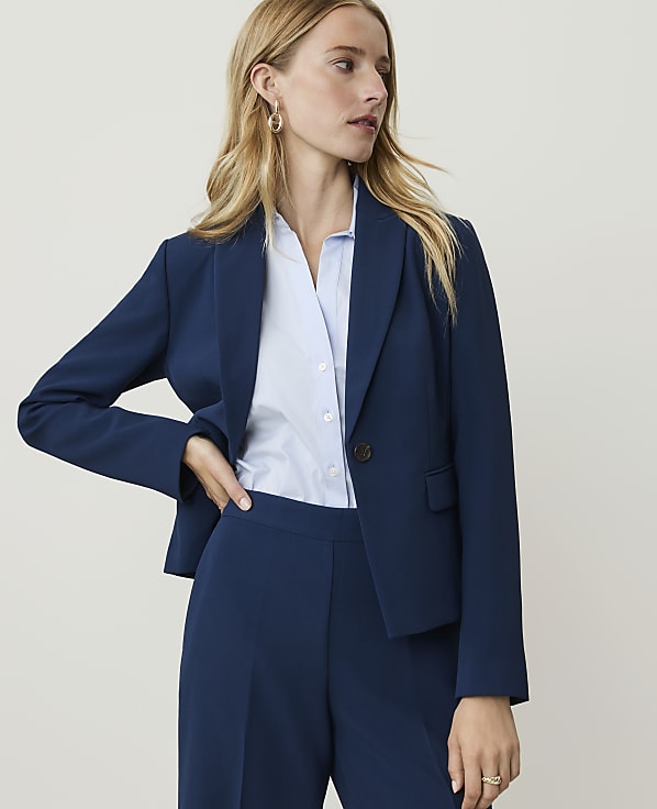A woman in a navy blue pantsuit stands against a plain, light background.