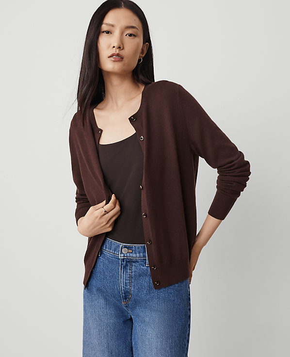 A woman in a brown cardigan and blue jeans stands against a plain white background.