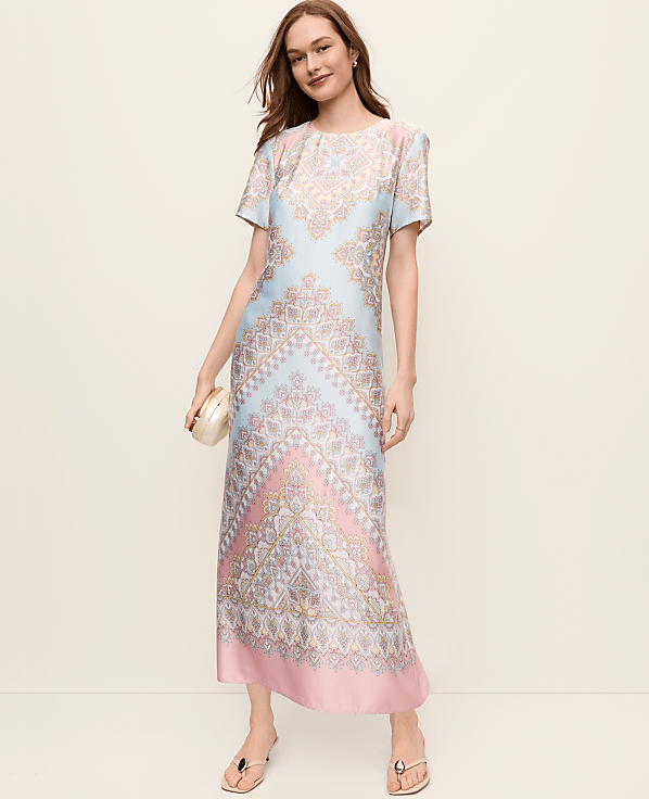 A young woman wearing a colorful, patterned dress stands in front of a plain, light-colored background, smiling confidently.