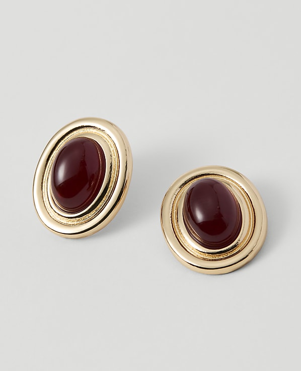 Two gold oval earrings with deep red cabochon stones are displayed against a plain, light gray background.