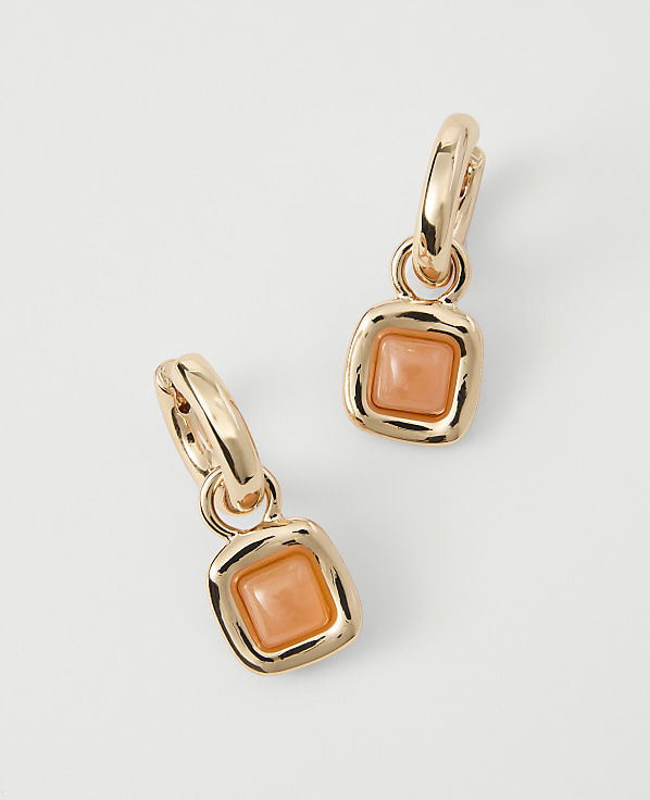 A pair of rose gold hoop earrings with square, peachy-orange stone pendants hang against a plain, light gray background.
