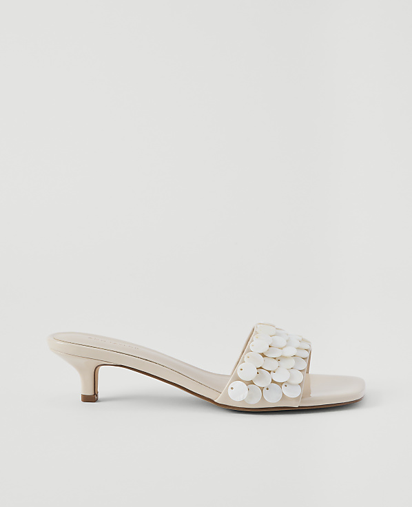 A cream-colored kitten heel sandal with a sequined strap rests on a plain, light gray background.