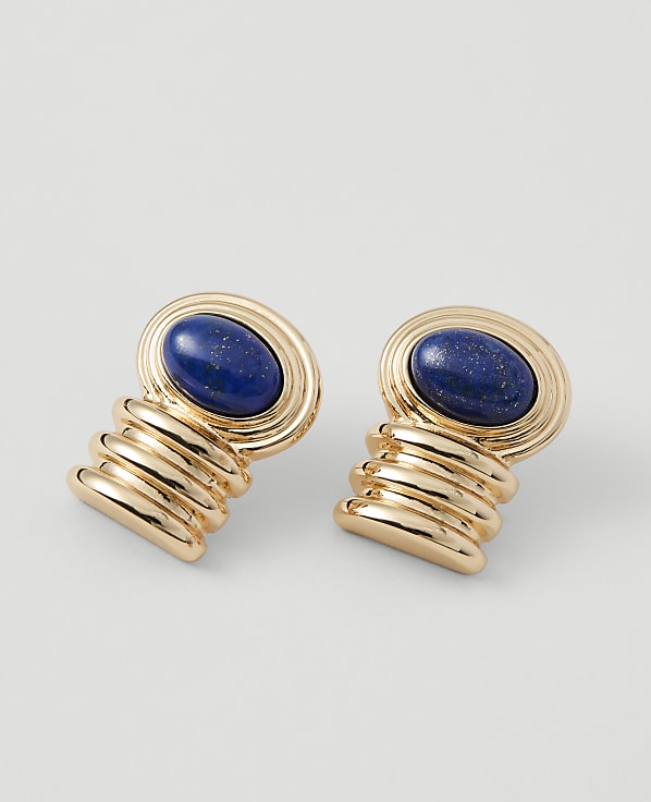 A pair of gold earrings with oval lapis lazuli stones are displayed against a plain, light gray background.