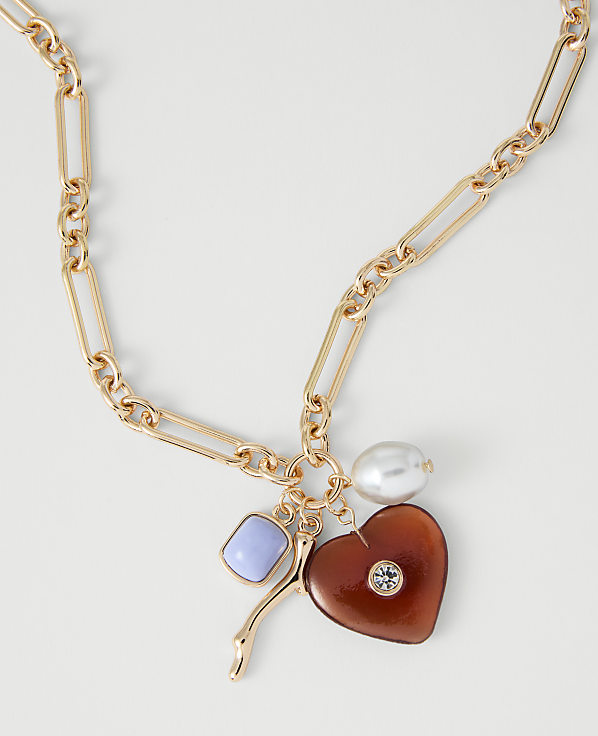 A gold chain necklace with a heart-shaped charm, a pearl, and a blue stone hangs against a plain white background.