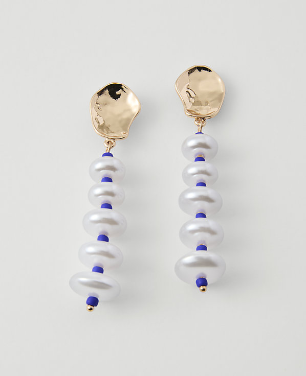 A pair of gold-topped earrings with stacked white pearls and blue beads hang against a plain white background.