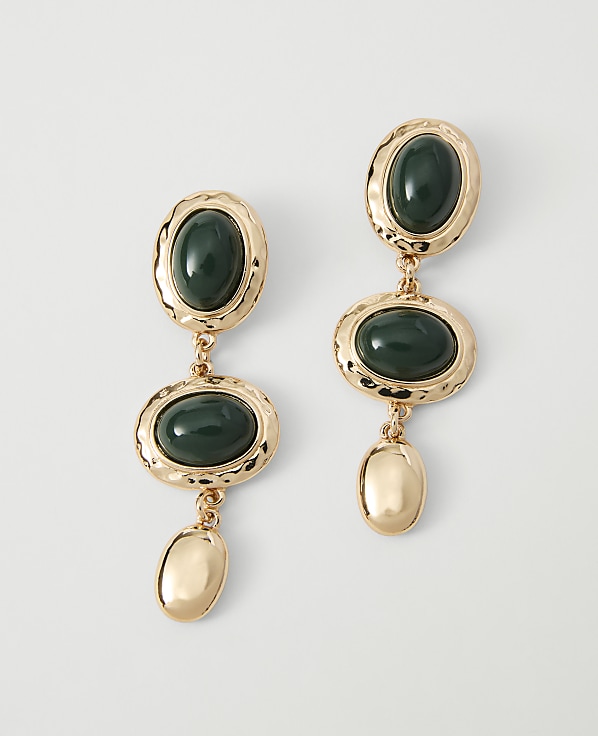 A pair of gold dangle earrings with oval green stones and gold accents hang against a plain white background.