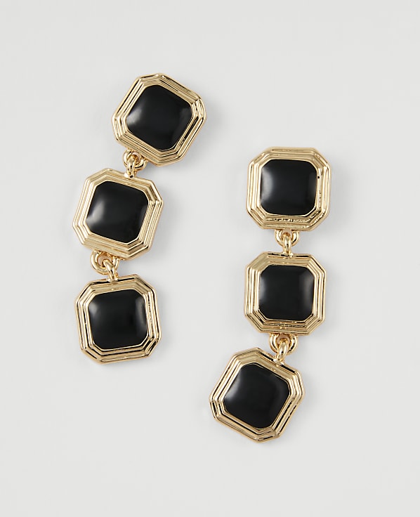 A pair of gold-toned dangle earrings with three black square charms each are laid out on a plain white background.