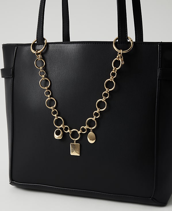 A black tote bag with a gold chain and charms hangs in the foreground against a plain white background.