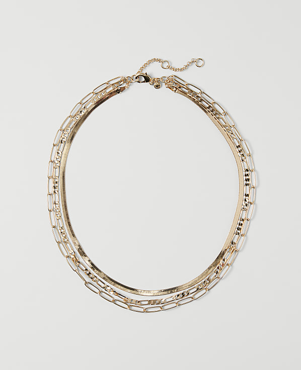 A layered gold necklace with a herringbone chain and rectangular links rests on a plain white background.