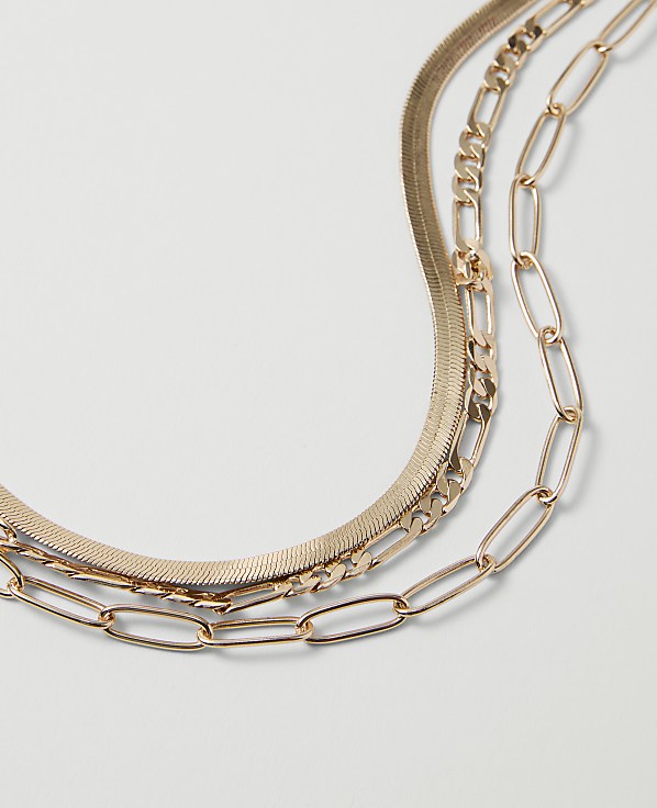 Three layered gold necklaces, a herringbone, a figaro, and a paperclip chain, rest on a plain white background.