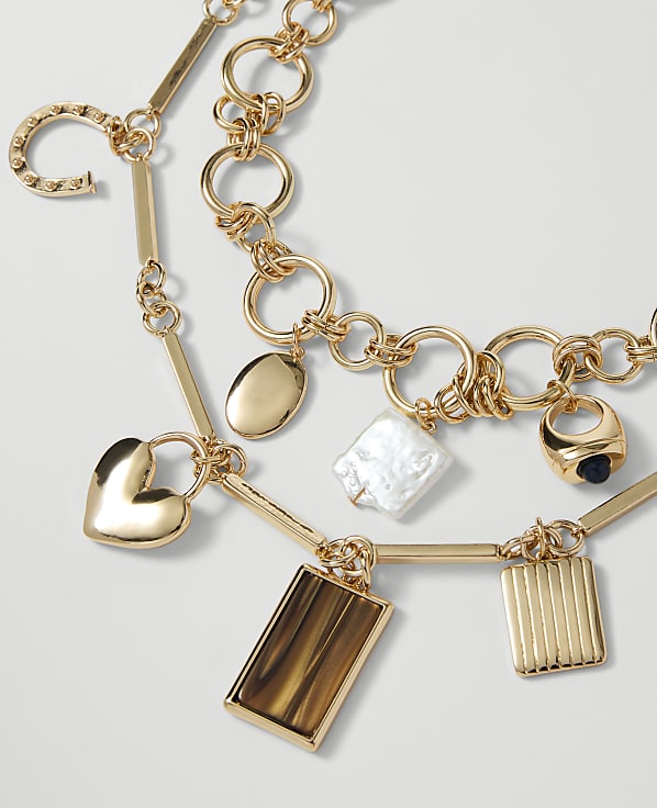 A gold necklace with various charms, including a heart, horseshoe, and rectangular pendants, rests against a plain white background.
