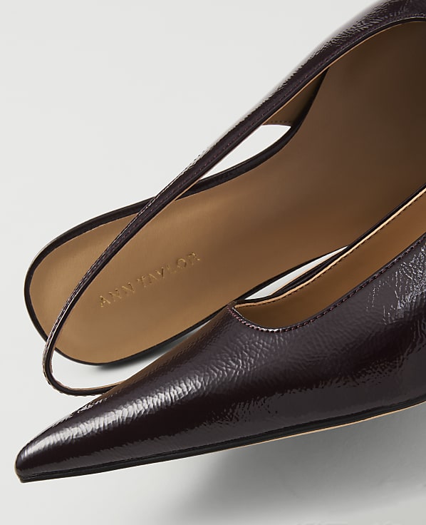 A close-up of a glossy, dark red pointed-toe pump with a thin strap is set against a plain white background.