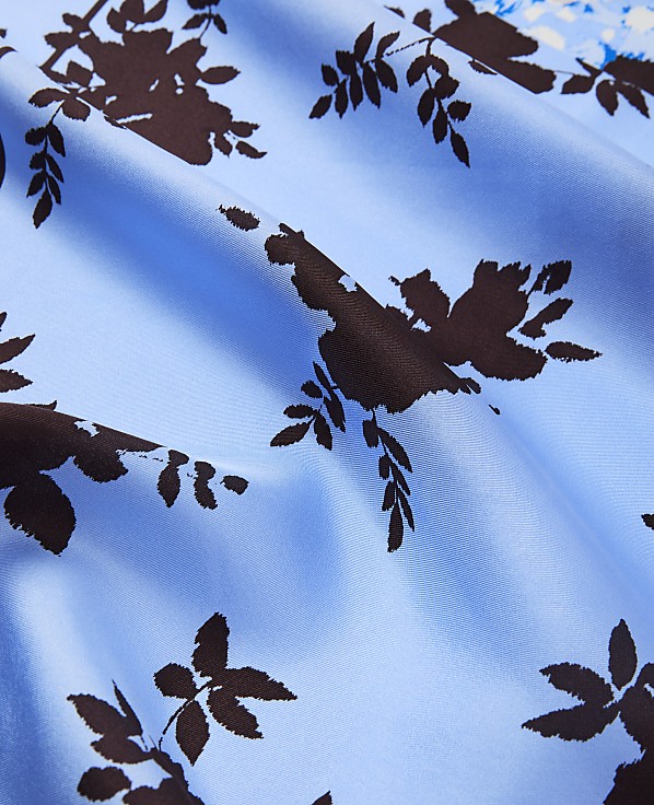 Brown floral patterns are scattered across a light blue, silky fabric in the foreground, with a hint of a lighter blue and white floral pattern visible in the blurred background.