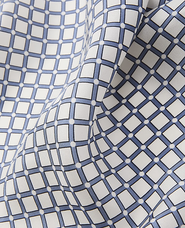 A close-up of white fabric with a repeating pattern of blue squares and white dots creates a textured foreground against a softly folded background of the same material.