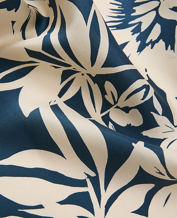 A close-up of cream-colored fabric with a bold, teal floral pattern fills the frame, with the fabric's folds creating depth and shadow.