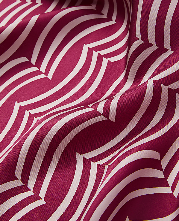 A close-up of a flowing, deep pink fabric with a repeating pattern of cream-colored, wavy lines creates a dynamic and textured foreground against a softly shadowed background.