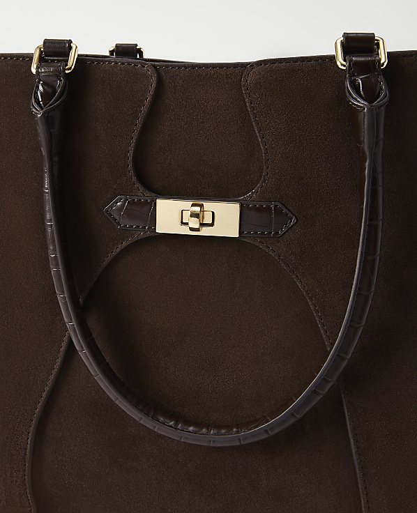 A close-up of a dark brown suede tote bag with a gold clasp and croc-embossed leather accents is presented against a plain white background.
