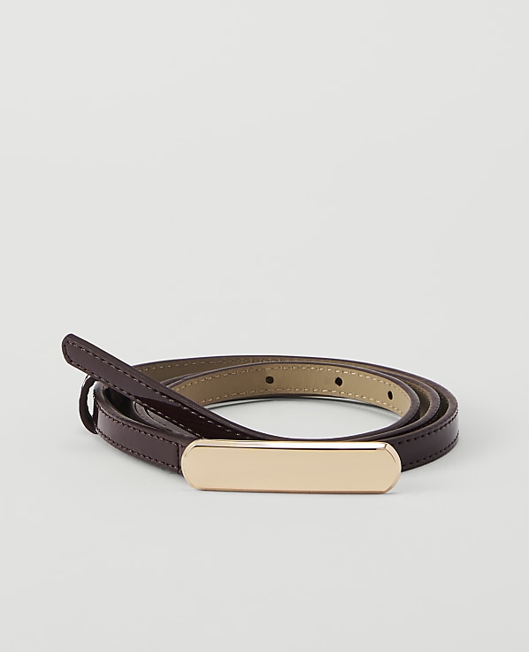 A thin, dark red patent leather belt with a gold buckle is coiled on a plain white background.