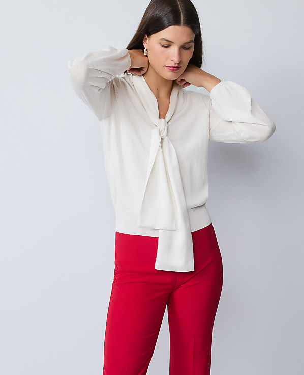 A woman in a cream bow-neck blouse and red pants stands against a plain gray background.