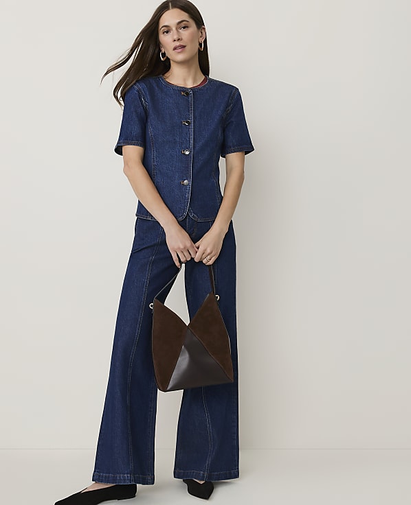 A woman in a denim outfit stands holding a brown handbag against a plain white background.