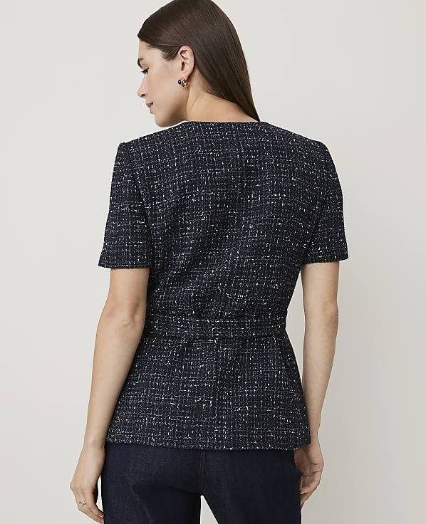 A woman in a dark tweed jacket with a cinched waist stands against a plain, light-colored background.