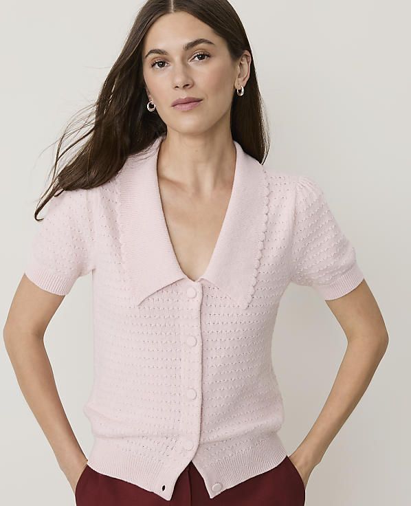 A woman in a pink collared cardigan stands against a plain white background.