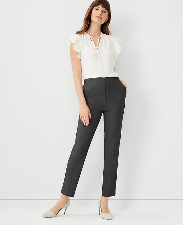 A woman in a white ruffled blouse and dark gray pants stands against a plain white background.