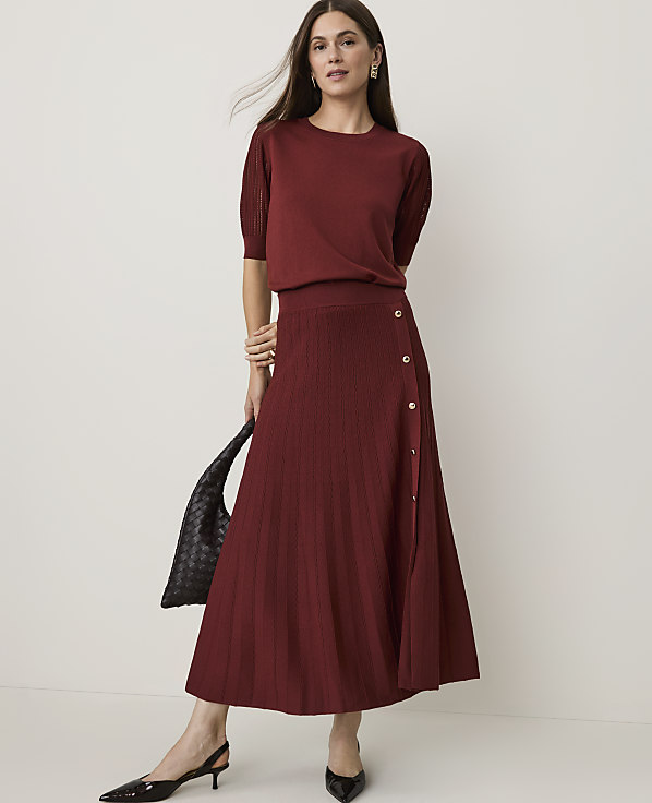 A woman in a burgundy knit set and black heels stands against a plain white background, holding a black woven bag.