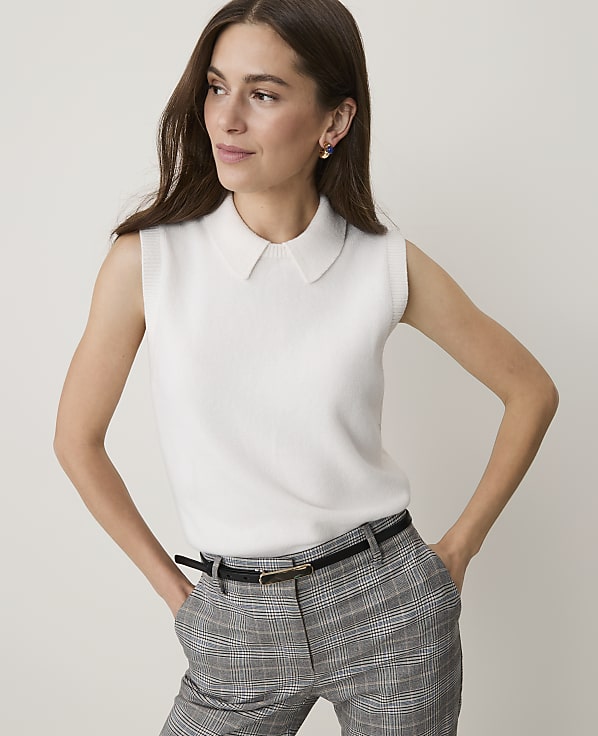 A woman in a white sleeveless collared top and plaid pants stands against a plain white background.