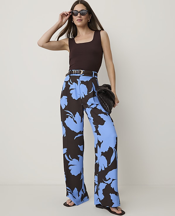 A woman in a brown tank top and blue floral pants stands against a plain white background, holding a dark clutch.