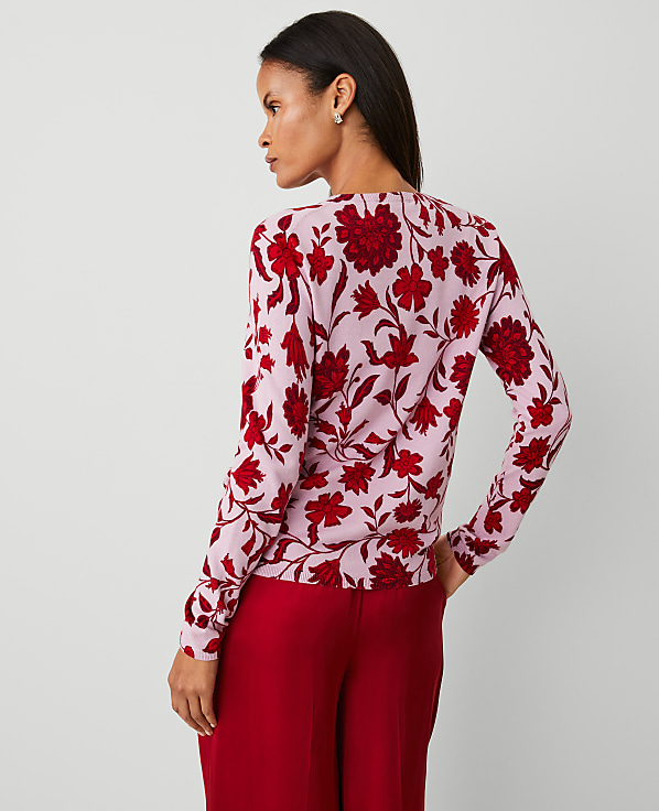 A woman in a pink floral sweater and red pants stands against a plain, light gray background.