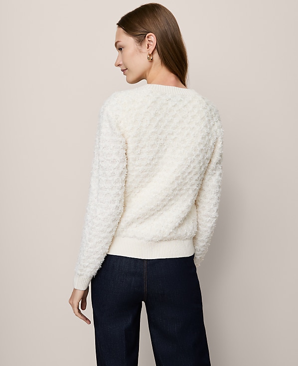 A woman in a textured cream sweater and dark jeans stands against a plain, light-colored background.