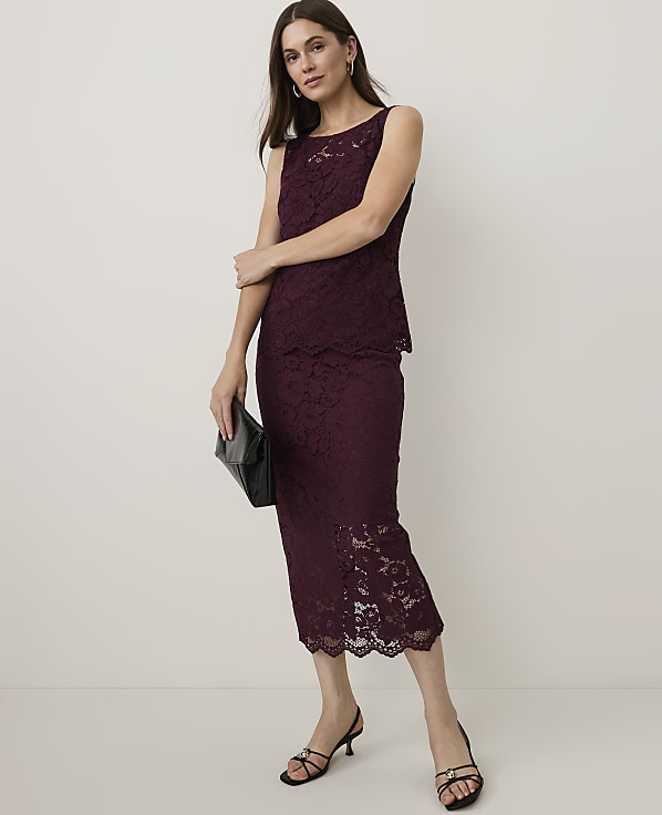 A woman in a burgundy lace dress and black heels stands with a clutch in front of a plain background.