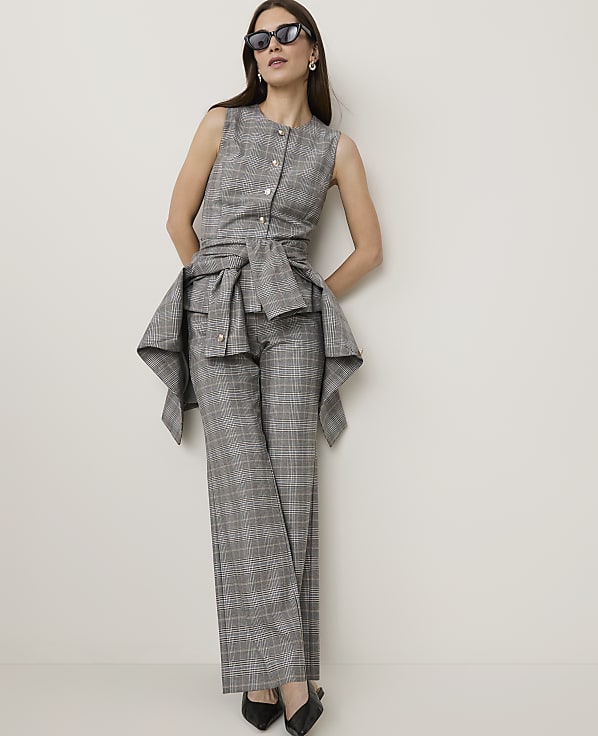A woman in a grey plaid suit with a peplum detail stands against a plain white wall.