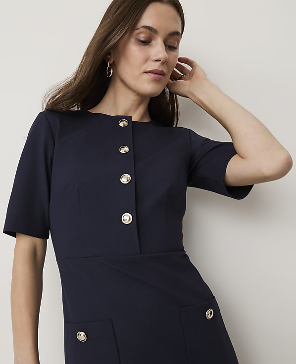 A woman in a navy dress with gold buttons stands against a plain white background.