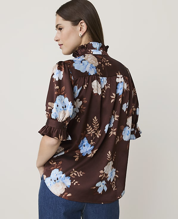 A woman in a brown floral blouse with ruffled sleeves and collar stands against a plain, light-colored background.